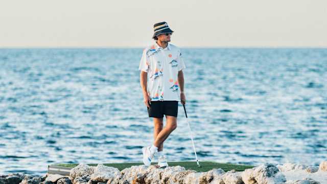 Rickie Fowler stands on a putting green near the ocean.